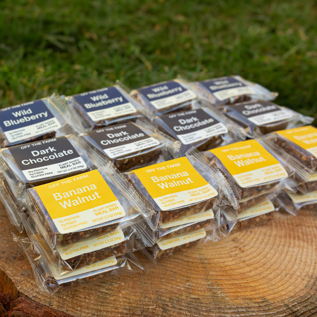 Assorted packaged OFF THE FARM meal bars on a wooden surface with grass in the background