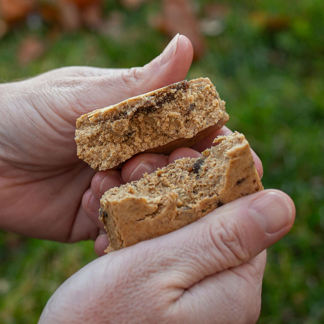 Hand holding two pieces of an OFF THE FARM PB Banana bar against a blurred outdoor background