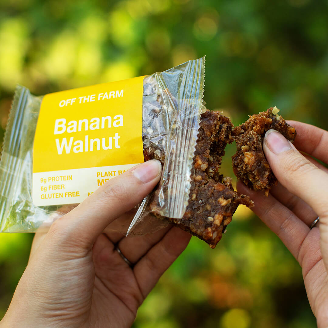 Hand holding a banana walnut meal bar with packaging against a blurred green background