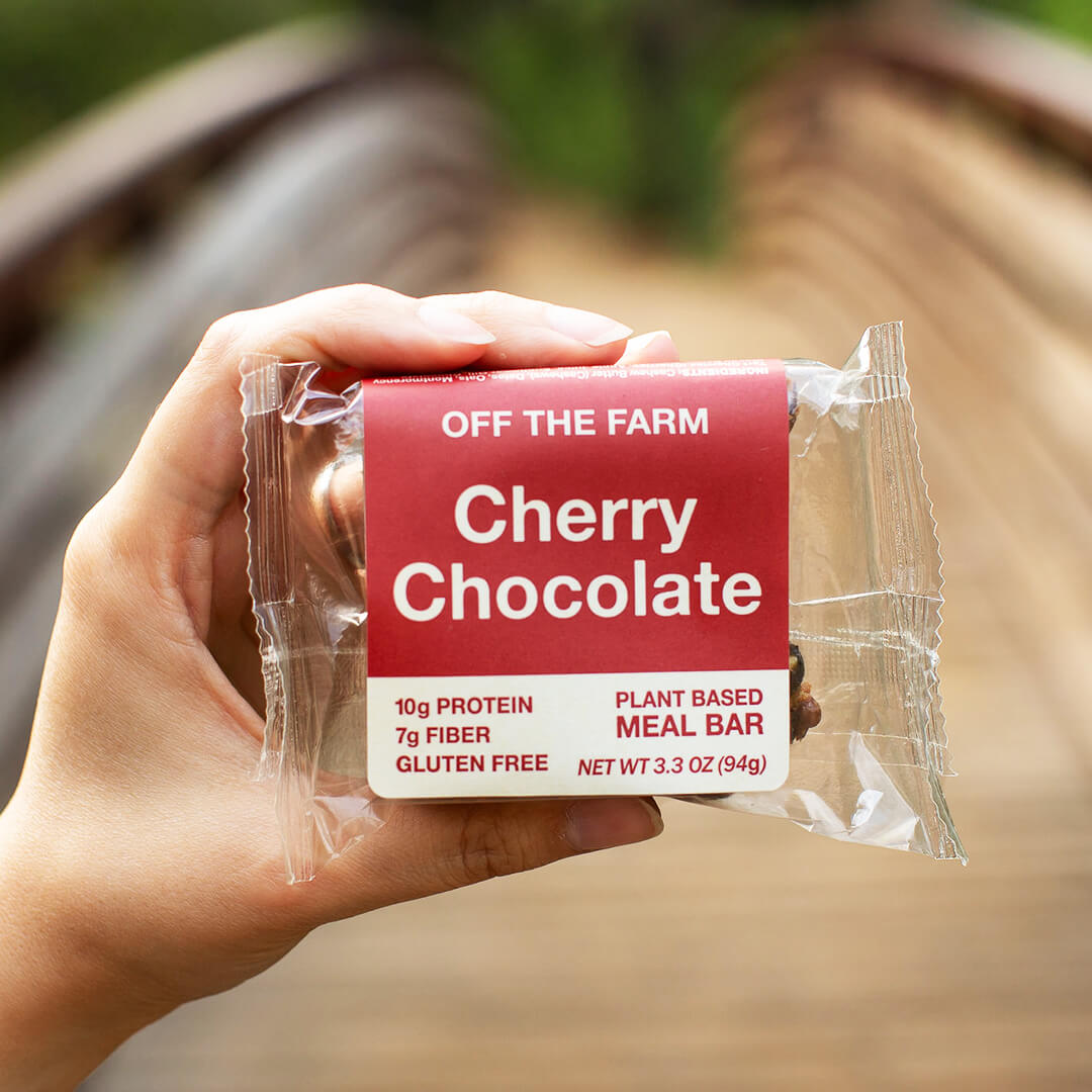 Hand holding a 'Off The Farm Cherry Chocolate' meal bar with a blurred natural background