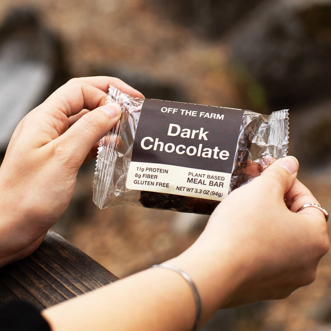 Person holding a package of 'Off The Farm' dark chocolate meal bar with a blurred natural background.