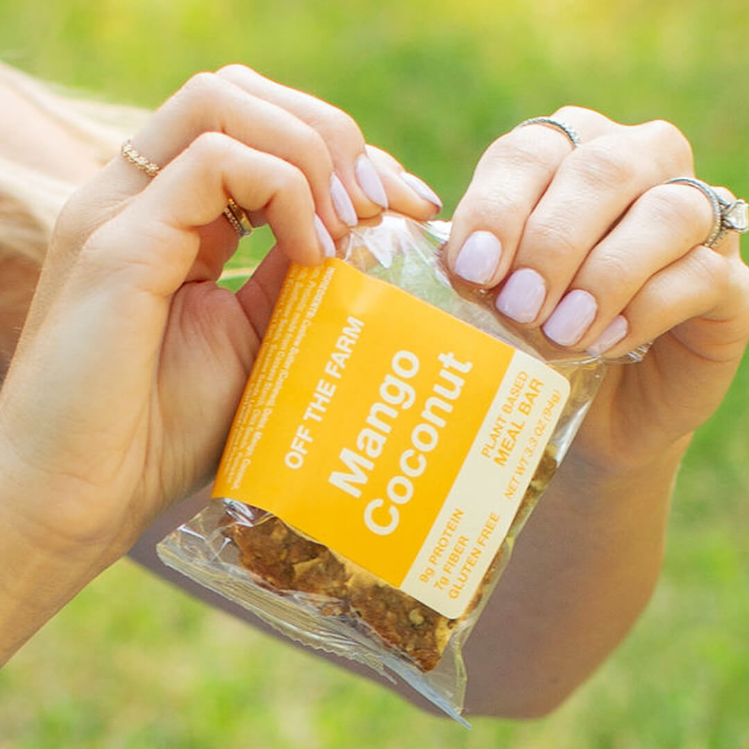 Hand holding a package of 'Off The Farm Mango Coconut' meal bar with a blurred green background