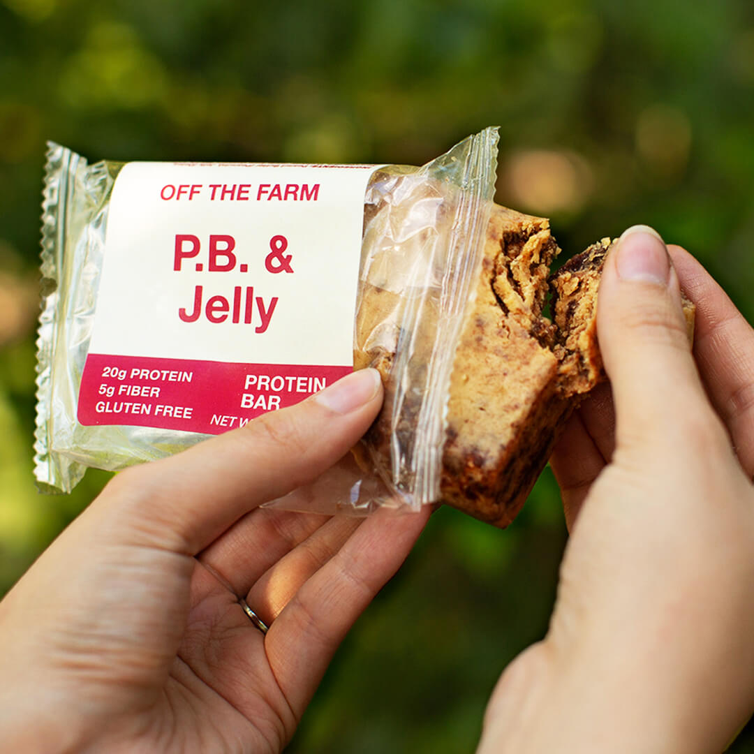 Hand holding a protein bar with 'P.B. & Jelly' packaging against a blurred green background