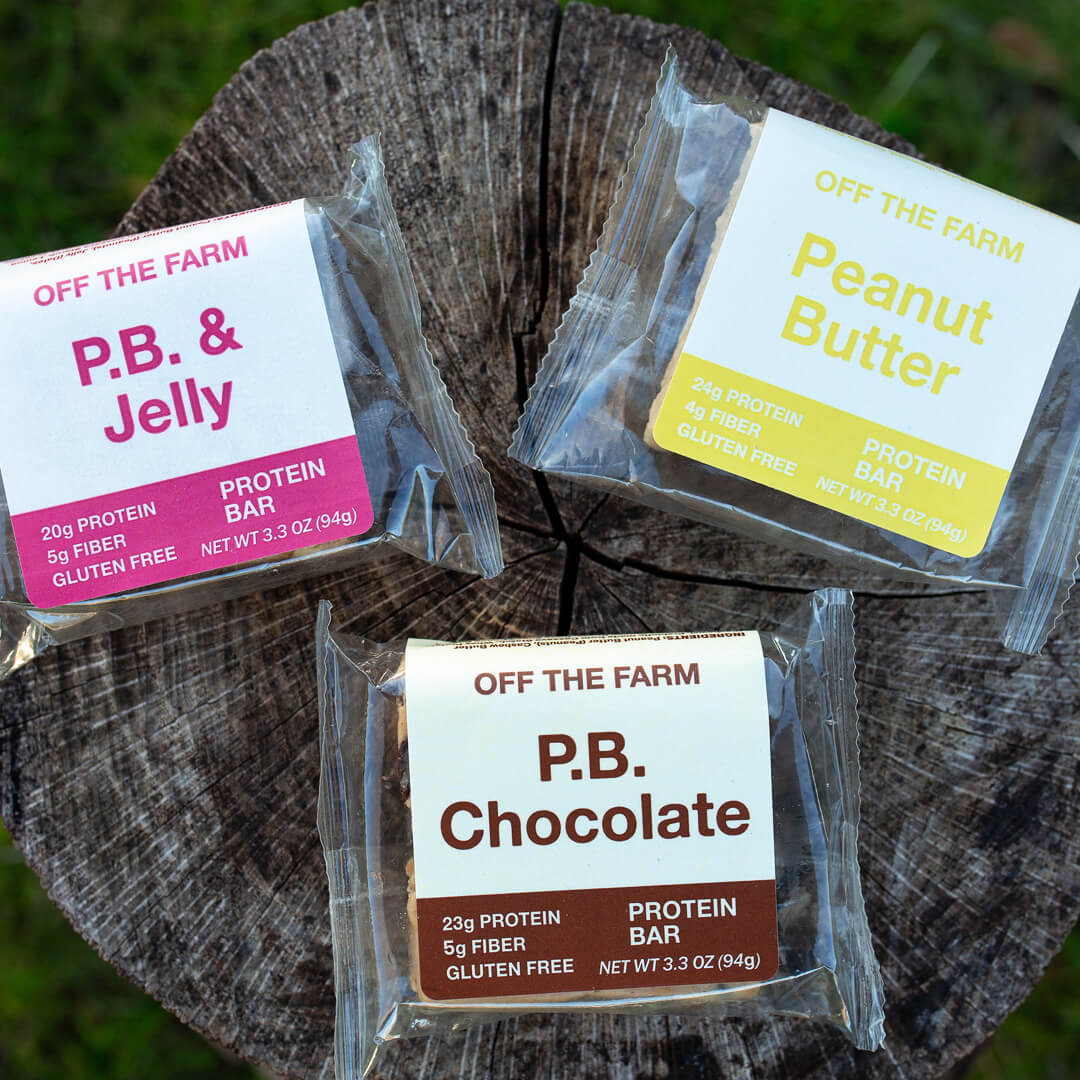 Three protein bars from 'Off The Farm' on a wooden stump with grass in the background.