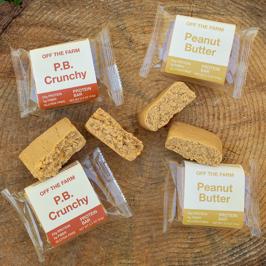 Four packaged protein bars from Off the Farm are arranged on a rustic wooden surface, with two peanut butter protein bars cut open to show the textured nut filling.