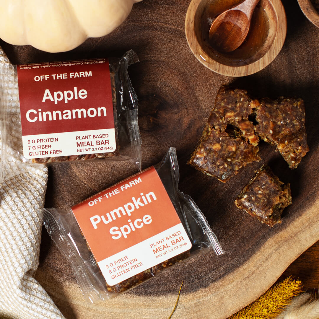 Two plant-based meal bars, one Apple Cinnamon and one Pumpkin Spice, on a wooden surface with a pumpkin and wooden bowl in the background.