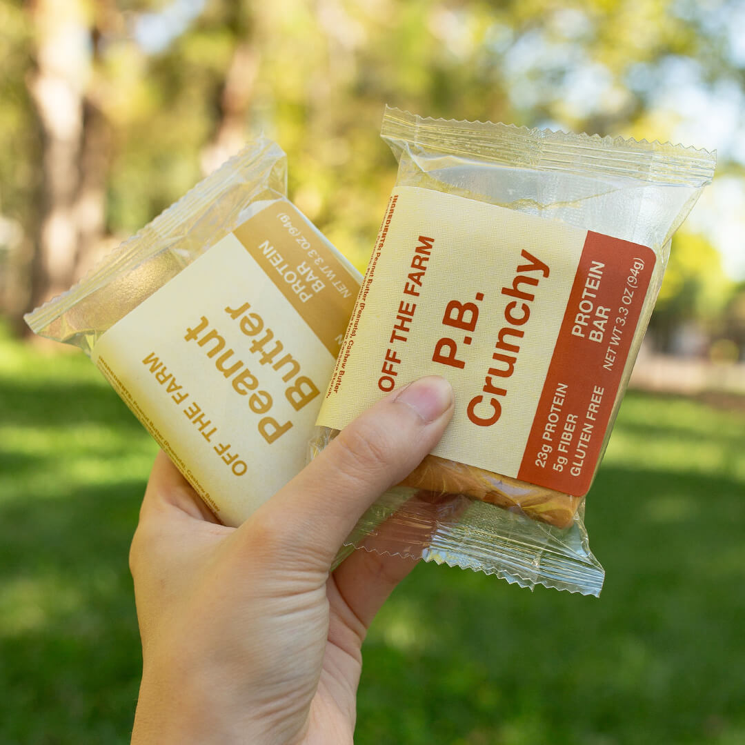 A hand holds two packaged protein bars outdoors in a park setting.