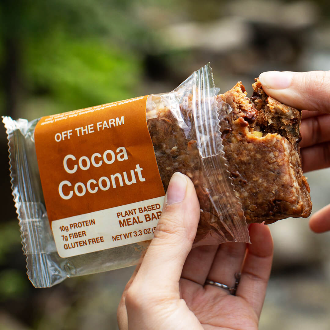 Hand holding a 'Cocoa Coconut' meal bar with 'Off The Farm' packaging against a blurred natural background.