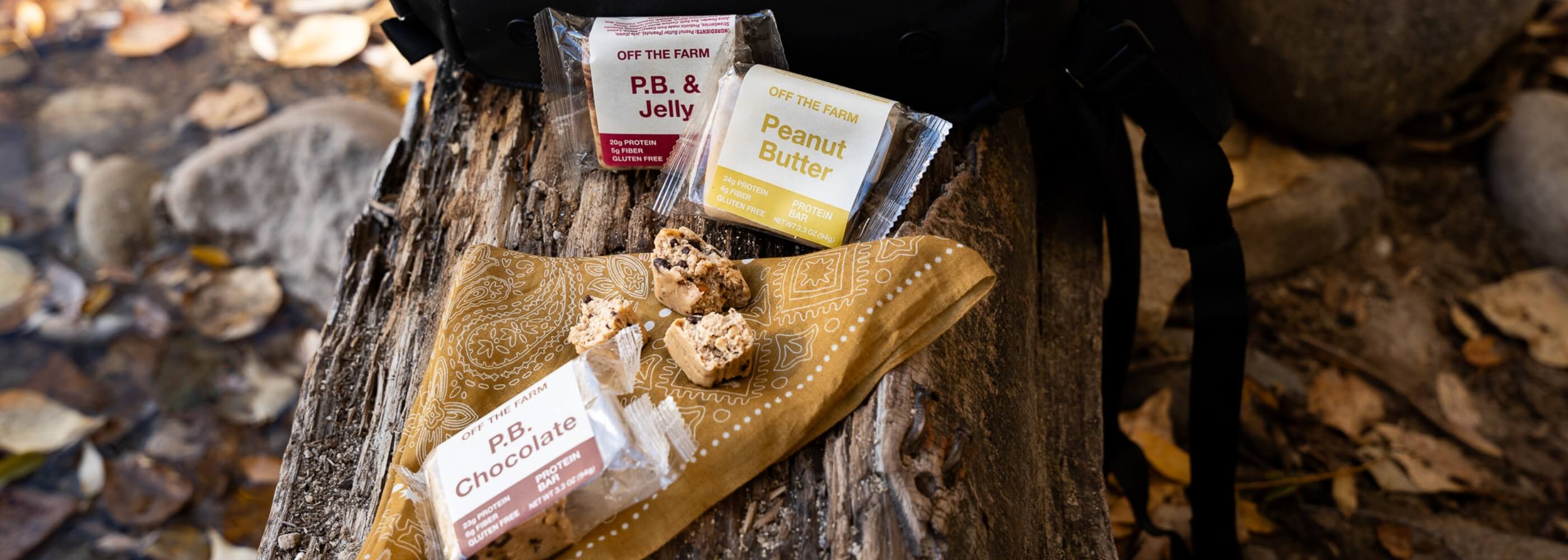Three Off The Farm protein bars sitting on a wood log on the ground outside.