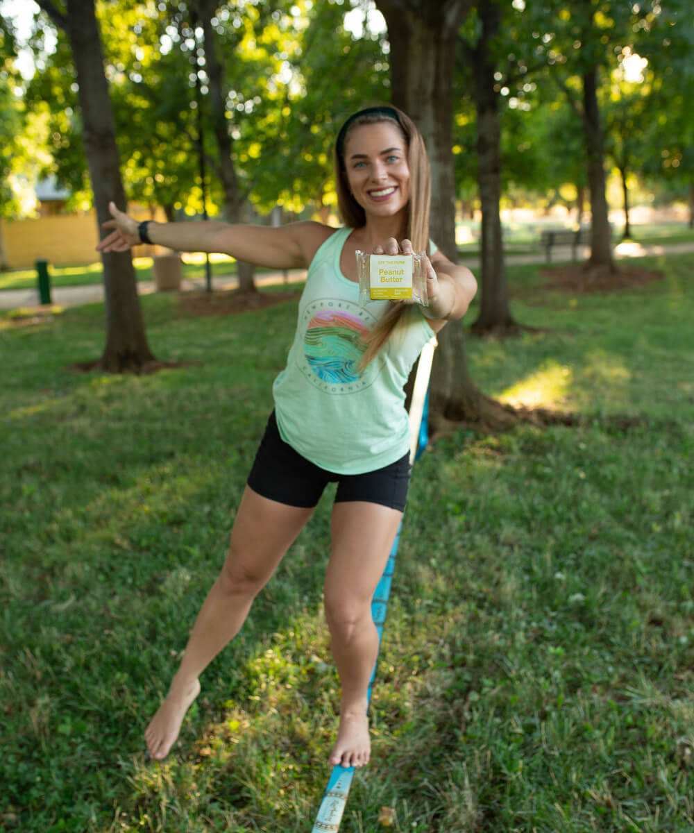 Woman in green shirt holding an OFF THE FARM peanut butter protein bar while balancing outside on a strip of cloth.