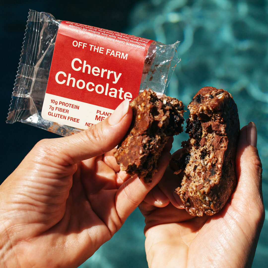 Hand holding a cherry chocolate snack bar with packaging labeled 'Off The Farm' against a blue background.