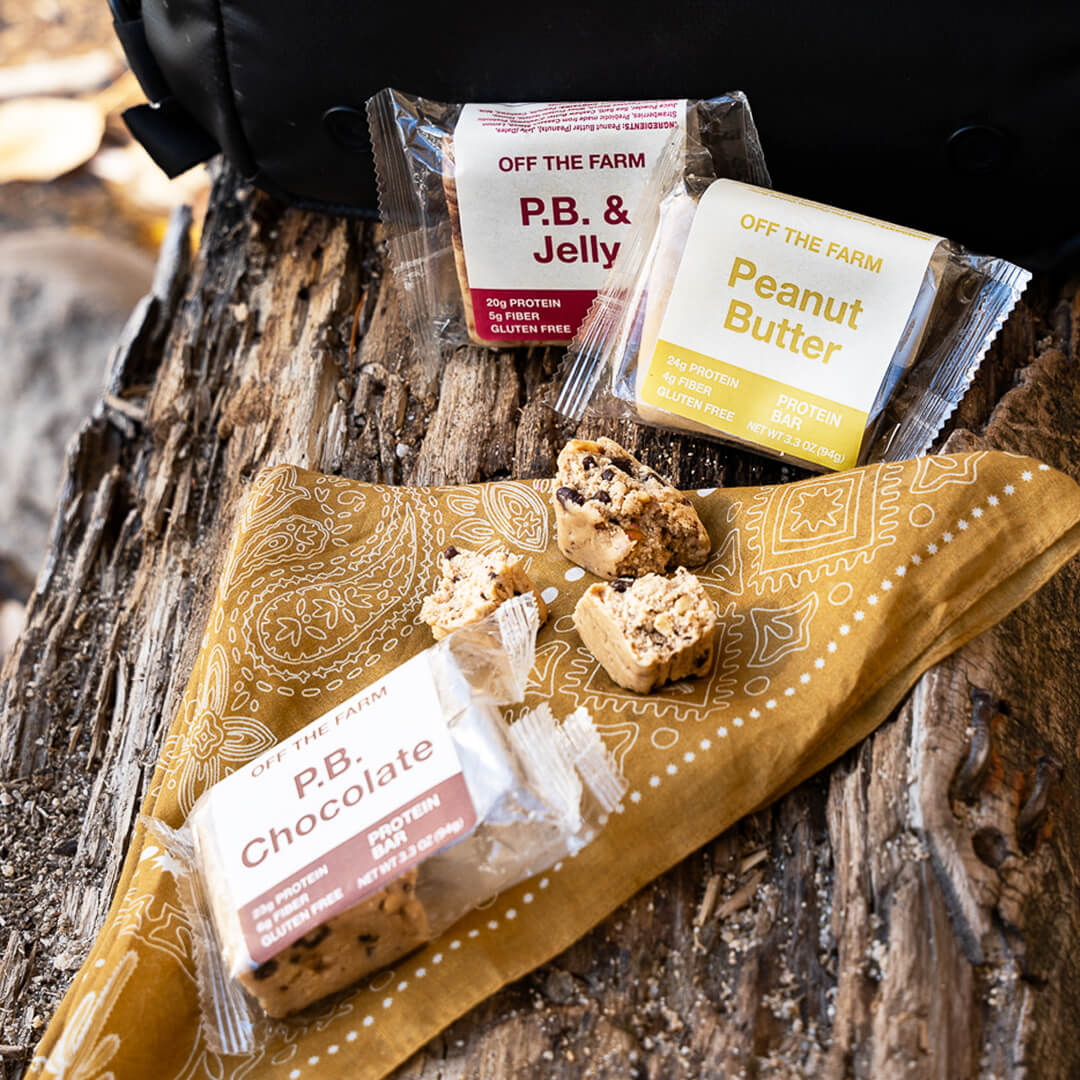 Peanut butter, P. B. & jelly, and P.B. chocolate protein bars on a wooden surface with a bandana.