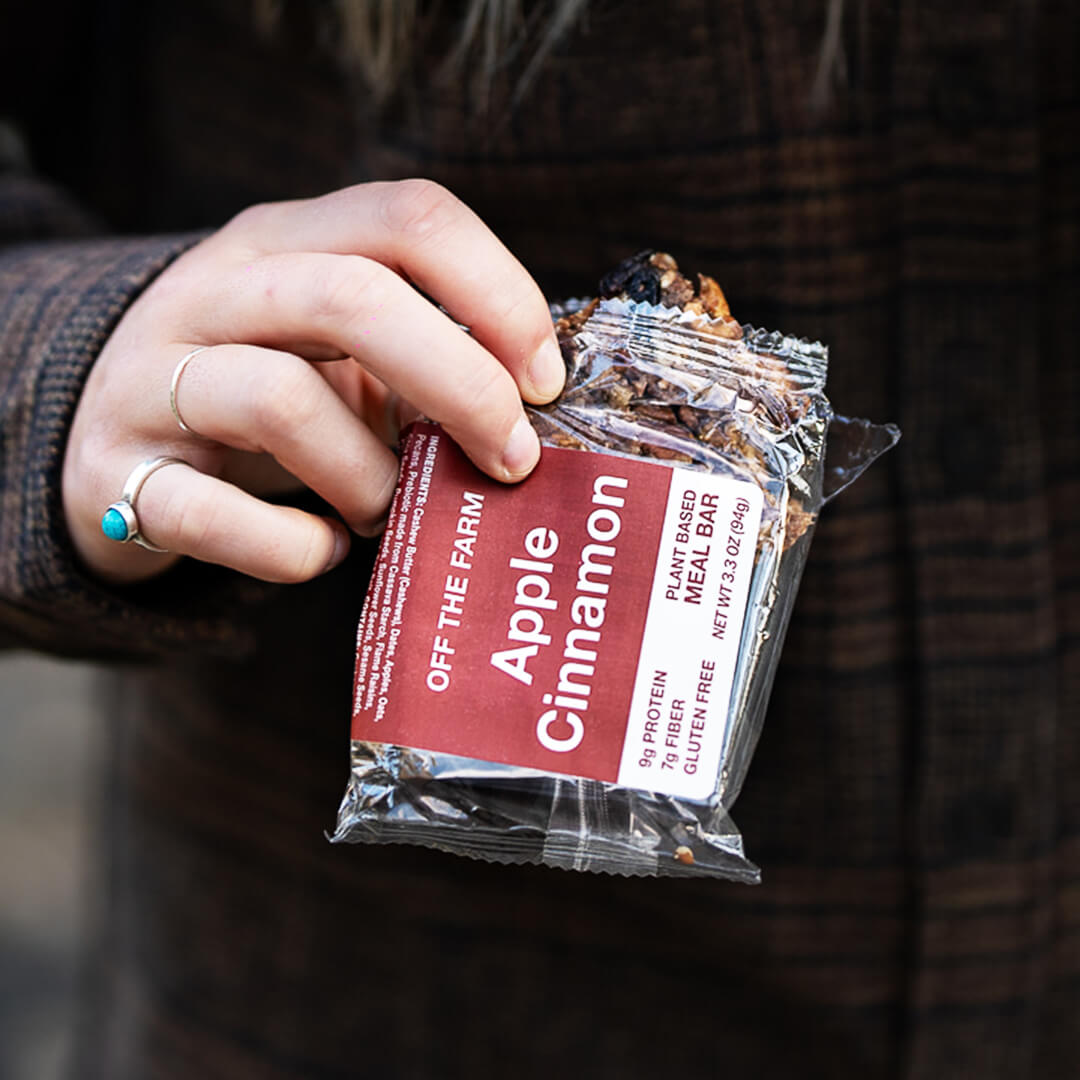 Hand holding a package of 'Off The Farm' Apple Cinnamon snack bar.