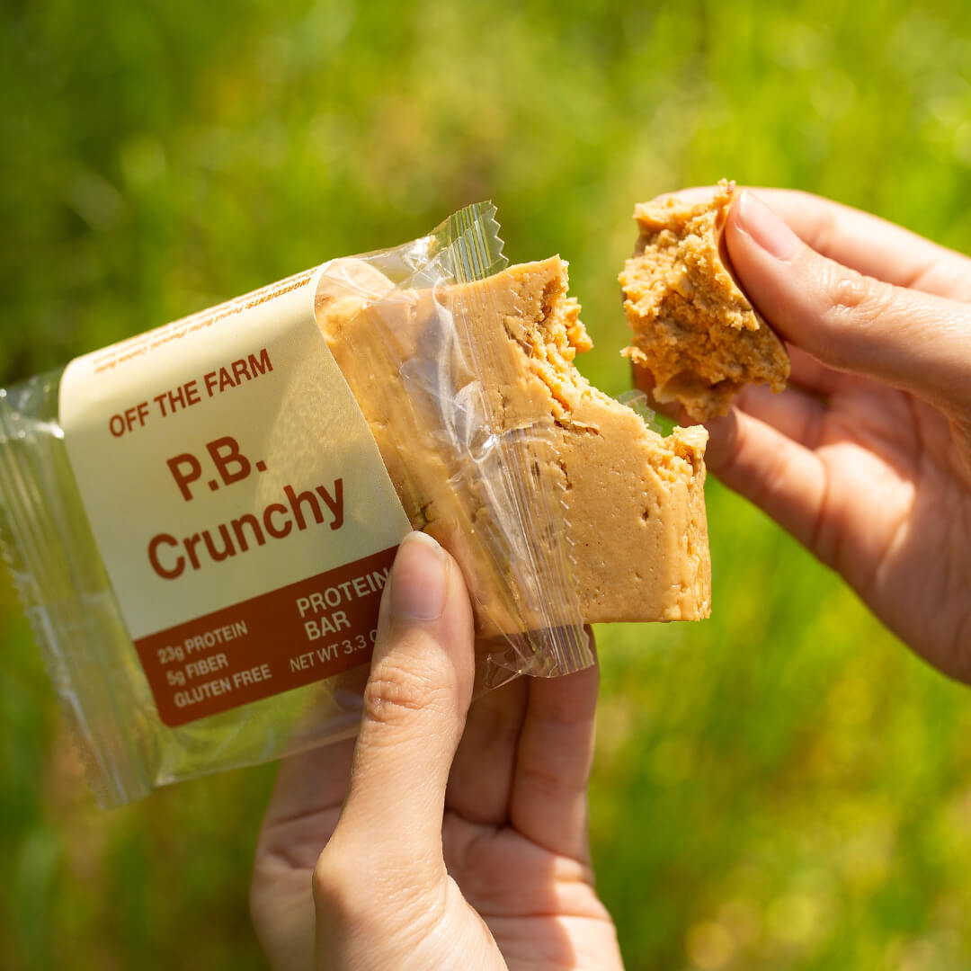 Person holding a P.B. Crunchy protein bar with a green outdoor background