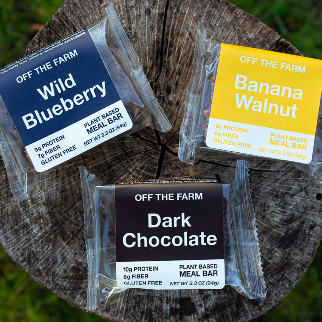 Three plant-based meal bar packages on a wooden surface, labeled 'Wild Blueberry', 'Banana Walnut', and 'Dark Chocolate'.
