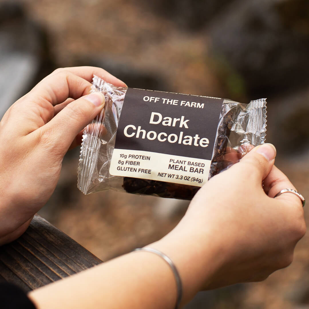 Person holding a package of 'Off The Farm' dark chocolate meal bar with a blurred natural background.