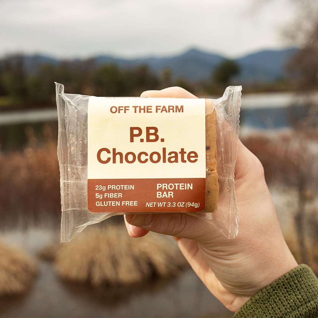 Hand holding a OFF THE FARM P.B. Chocolate protein bar with a scenic background