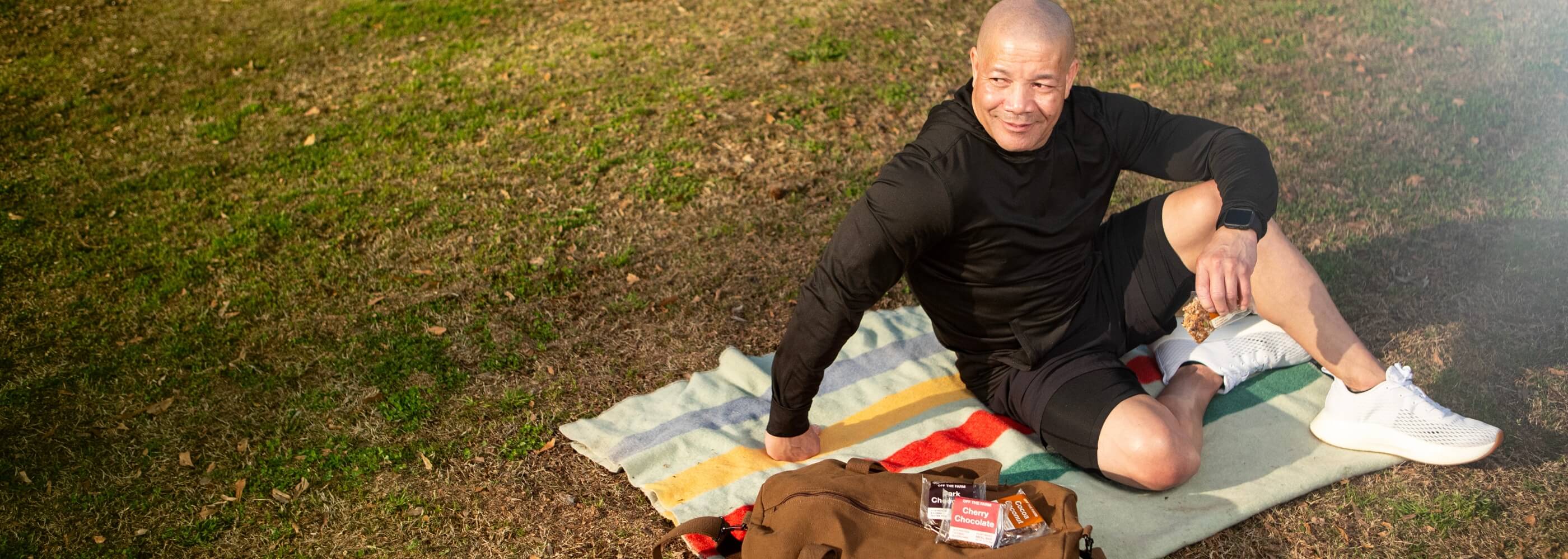 A person sits on a blanket in a grassy area, wearing athletic clothing and white shoes, enjoying a snack. A bag and snacks are nearby.