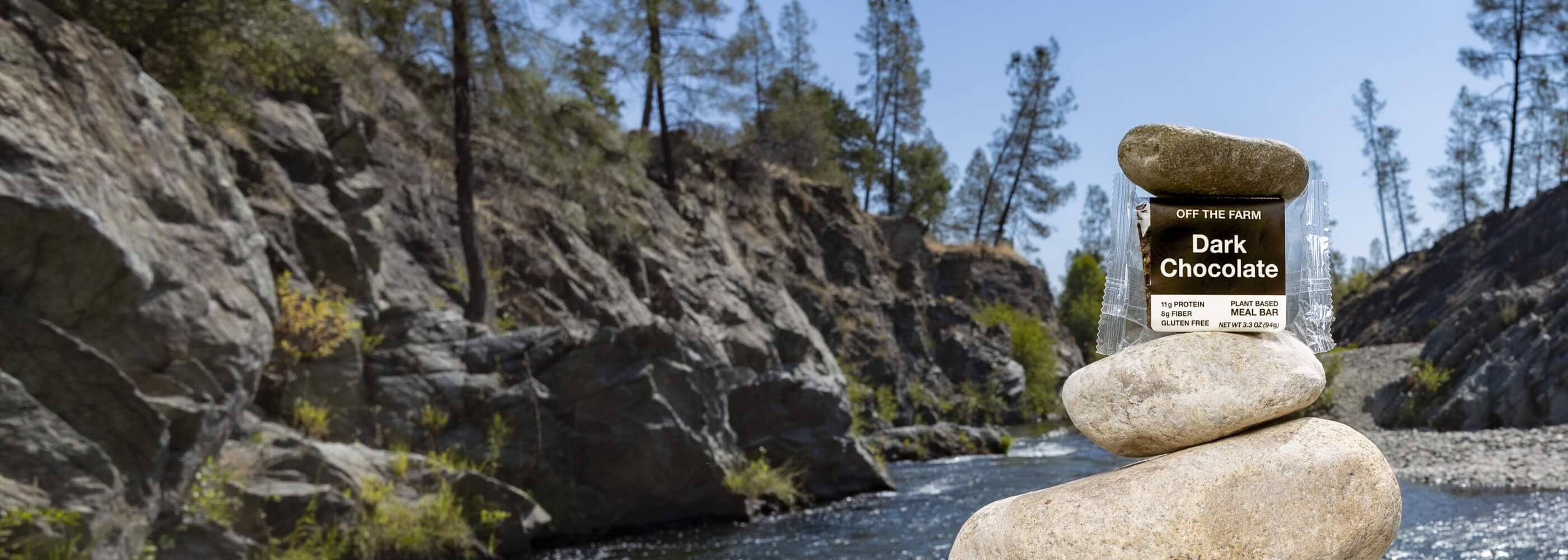 Off The Farm Dark chocolate meal bar sitting between two rocks by a river.