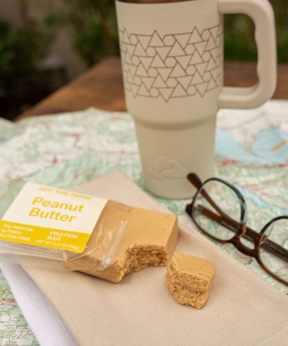 OFF THE FARM peanut butter protein bar on napkin next to glasses, a map and a coffee mug, all sitting on a table